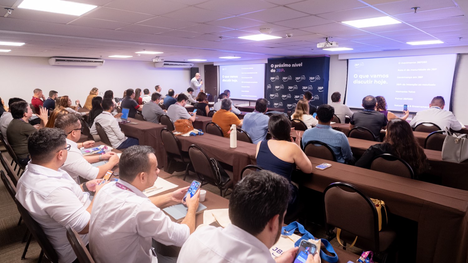 Sala de eventos em Fortaleza com cerca de trinta participantes sentados em mesas dispostas em U, voltados para dois telões com a abertura do JBP360 e a agenda do dia.