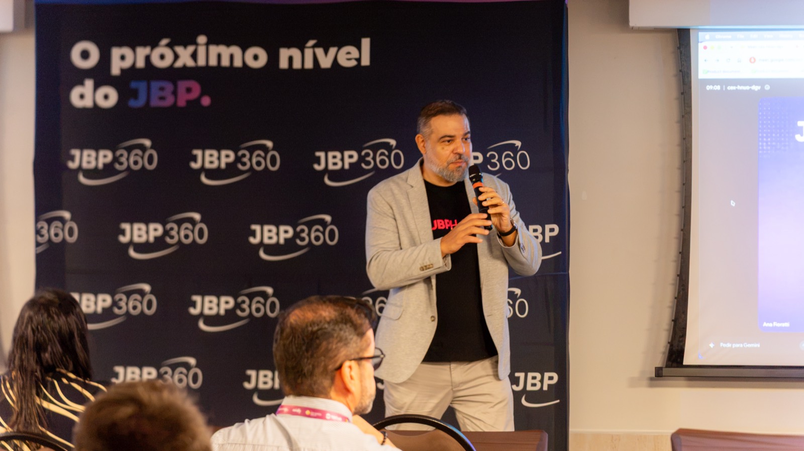 Guilherme Auger, da Midfy, fala ao microfone diante de um painel preto com logotipos JBP360 repetidos e a frase O próximo nível do JBP.