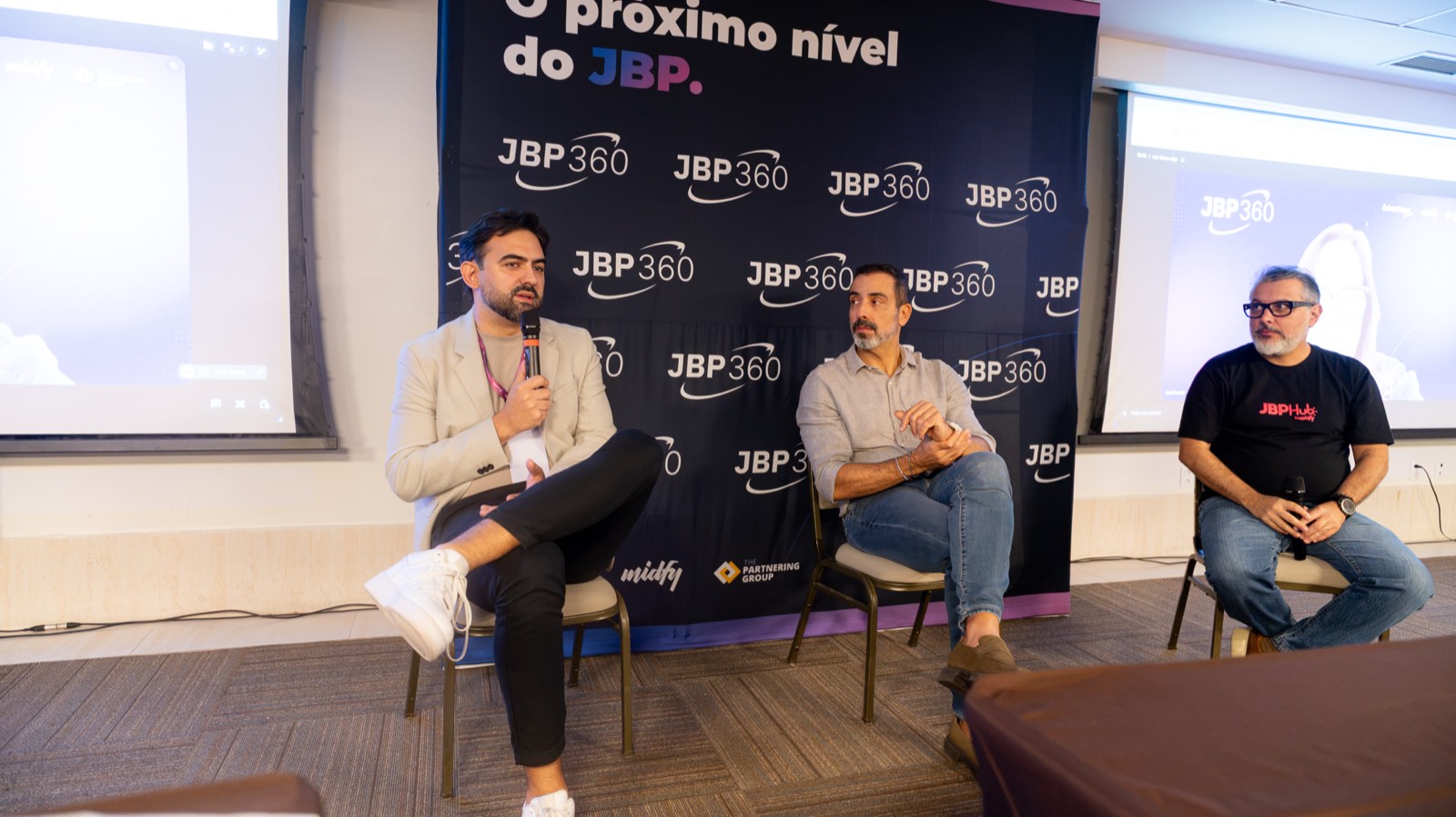 Dois painelistas sentados lado a lado diante do painel de fundo do JBP360; um deles fala ao microfone e gesticula.
