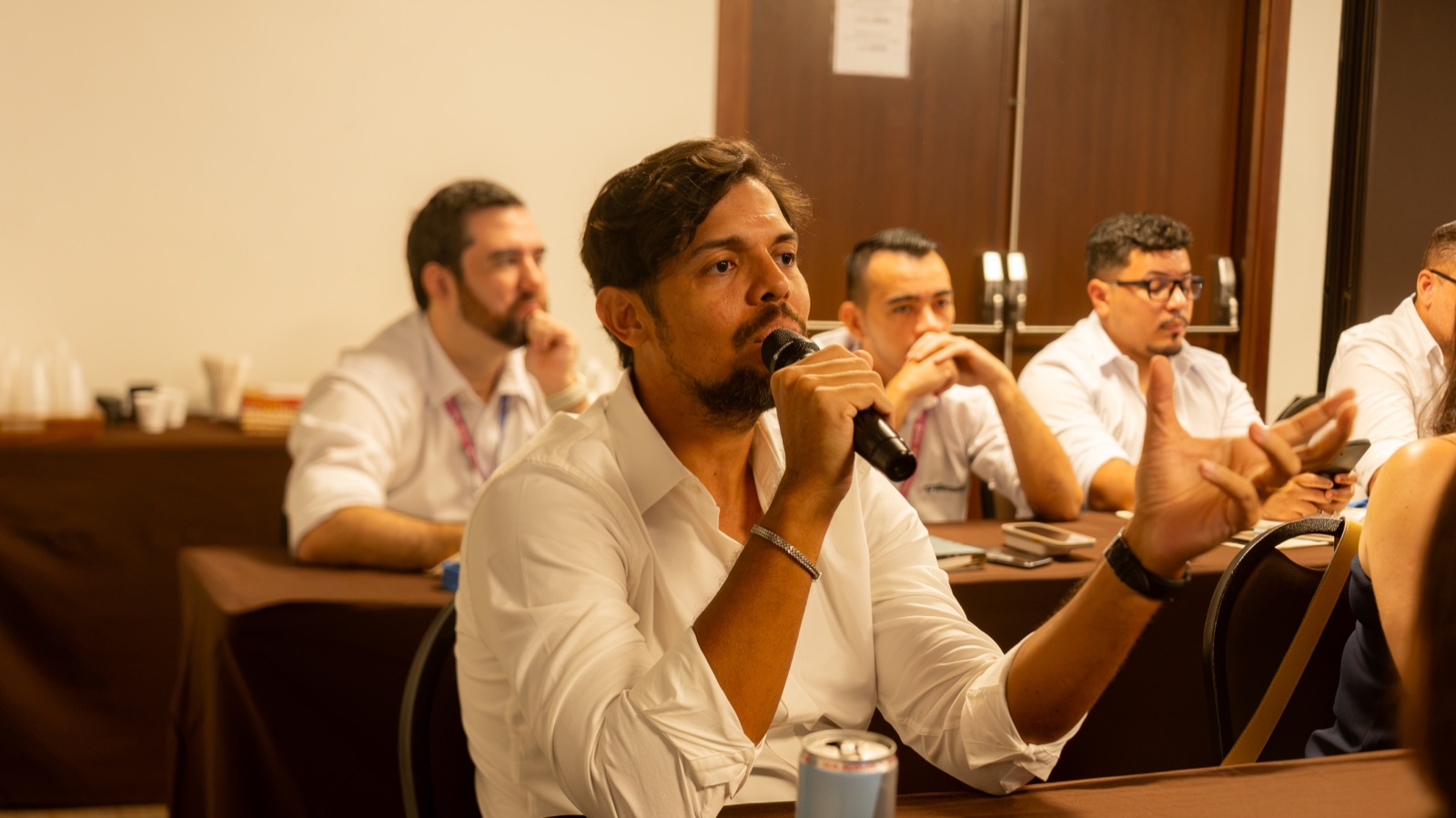 Participante de camisa branca faz uma pergunta ao microfone durante o encontro, com outros participantes atentos ao fundo.