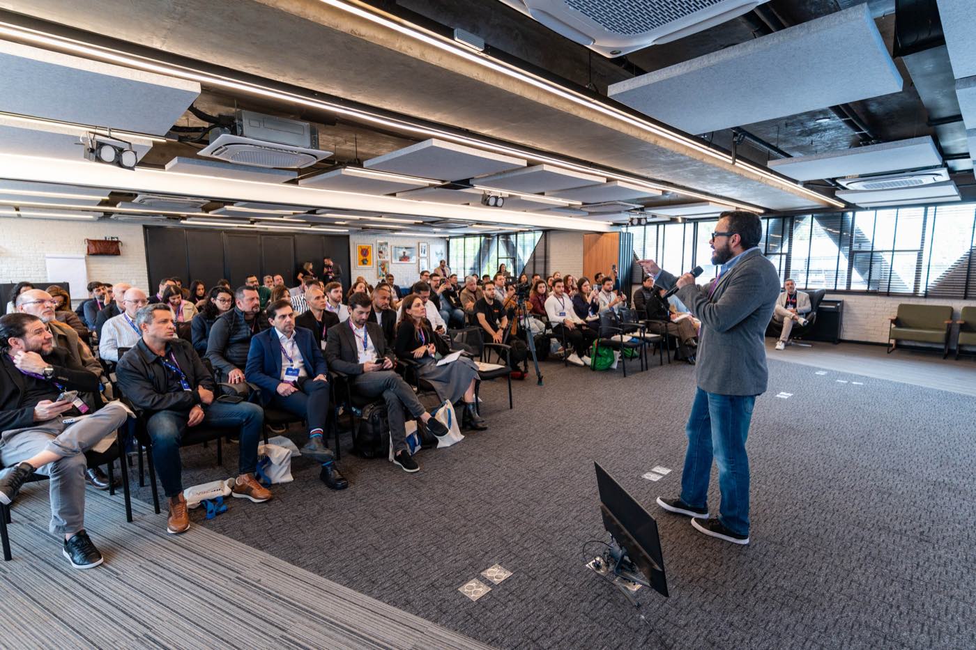 Sala do Dinastia Hub cheia, com dezenas de participantes sentados em cadeiras enfileiradas e um palestrante em pé ao microfone à direita.