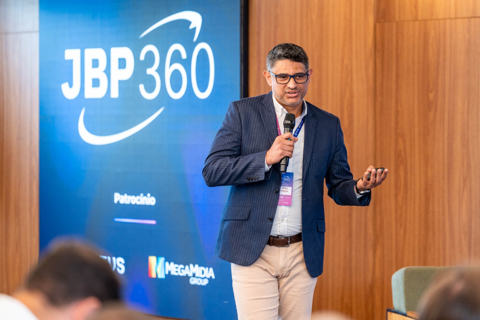 Guilherme Auger, da Midfy, fala ao microfone diante de um telão azul com a marca JBP360 e os logotipos dos patrocinadores Totvs e MegaMidia.