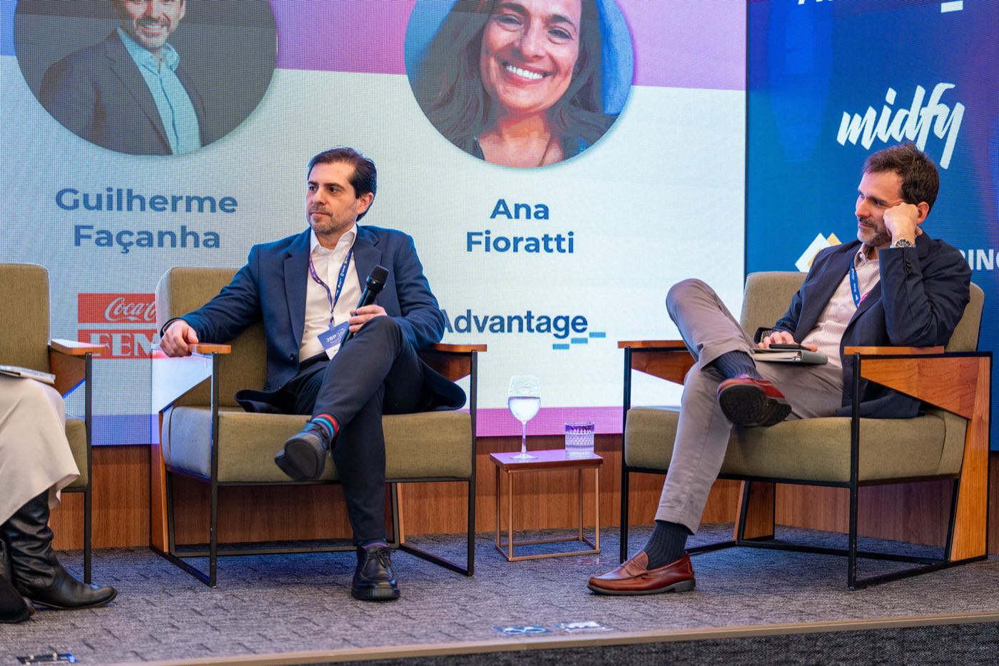 Painelistas sentados em poltronas no palco durante o painel; ao fundo, o telão exibe as fotos e os nomes dos participantes da conversa.