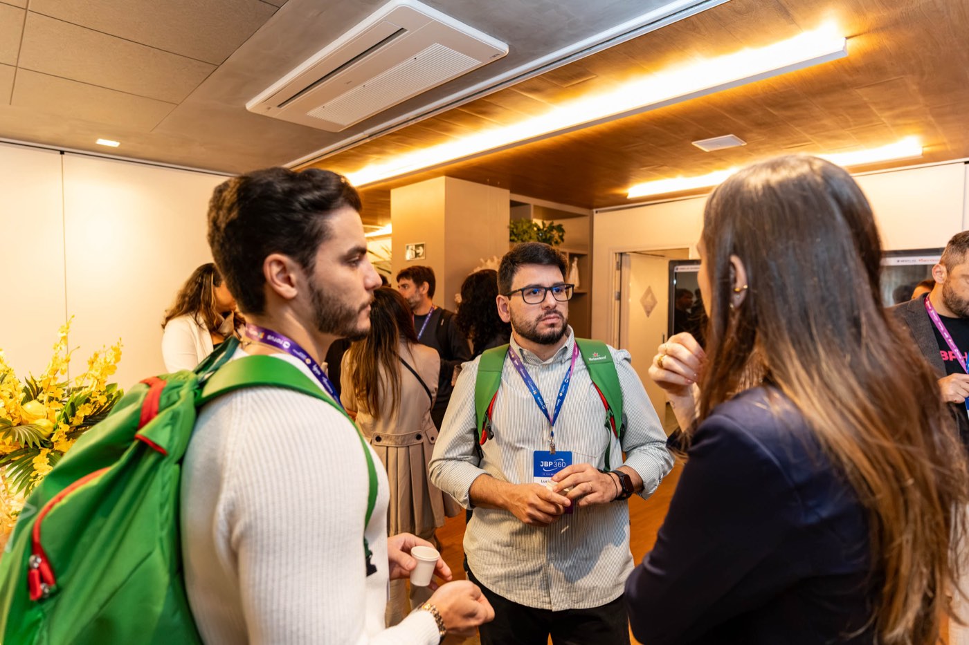 Três participantes com crachás do JBP360 conversam em pé no hall do evento durante o intervalo.